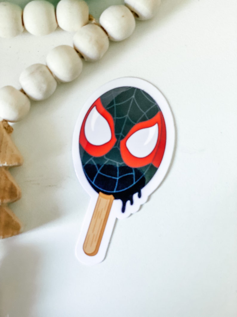 May include: A black and red Spider-Man face on a white popsicle stick. The popsicle is dripping black and has a white background.