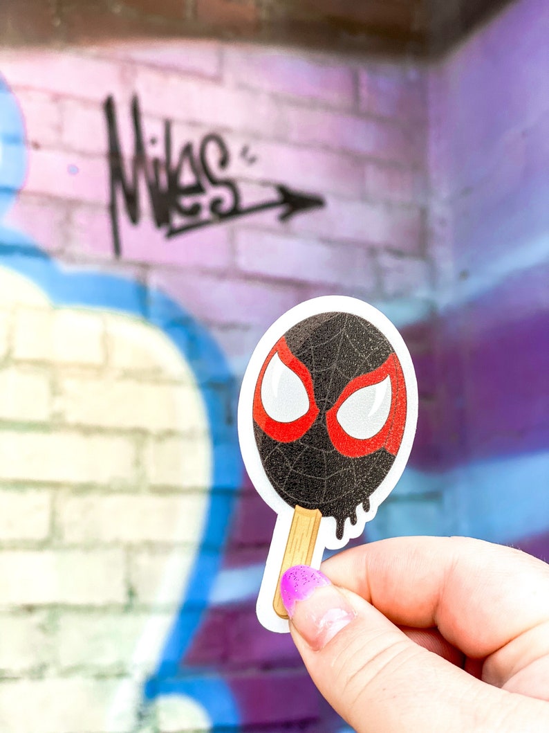 May include: A black and red Spider-Man popsicle sticker with a white background. The sticker is shaped like a popsicle with a wooden stick.