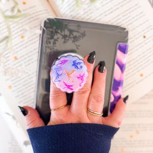 May include: A round phone grip featuring a dragon design in shades of pink, purple, and blue. The grip is attached to a black phone case. The background includes a book and a navy blue sleeve.