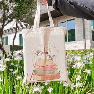 May include: A beige tote bag with a design of a stack of books with the words "Dragon Fandom", "Wizard Fandom", and "Fae Fandom" on the spines. The bag also has a design of a sword and two dragons with the text "I was enchanted to meet you".