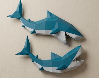 May include: Two geometric paper shark wall decorations. The sharks are teal and white, with open mouths revealing white teeth. The paper sharks are mounted on a neutral-colored wall.