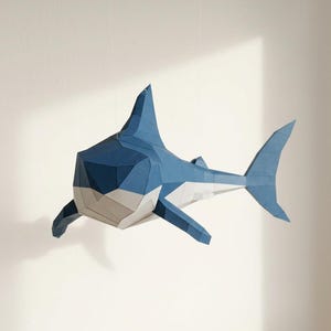 Op de afbeelding: Een geometrische papieren haai sculptuur in tinten blauw, wit en grijs, opgehangen tegen een witte achtergrond. De haai heeft een gedetailleerd, hoekig ontwerp, met vinnen en een staart. Het kunstwerk is een creatieve weergave van het zeeleven.