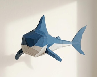 May include: A geometric paper shark sculpture in shades of blue, white, and gray, suspended against a white background. The shark features a detailed, angular design, with fins and a tail. The artwork is a creative representation of marine life.