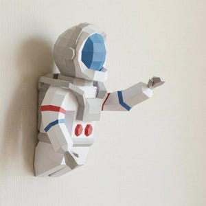 May include: A white and gray astronaut wall decoration with red and blue accents. The astronaut is reaching out with one arm. The helmet has a blue visor. The design is geometric and three-dimensional.