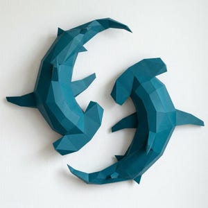 以下が含まれることがあります： 2つのティール色の幾何学的なシュモクザメのウォールアート。サメは折り紙または厚紙で作られており、鋭い角度と多角形のデザインが特徴です。サメは白い背景に表示されています。