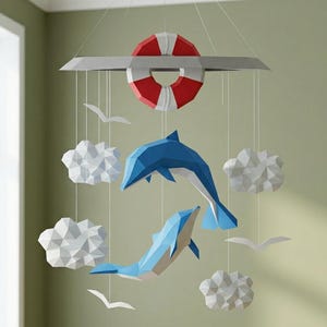 Puede incluir: Móvil de temática náutica con un salvavidas rojo y blanco, dos delfines azules, nubes blancas y gaviotas. El móvil está suspendido de una barra gris, ideal para una habitación infantil.
