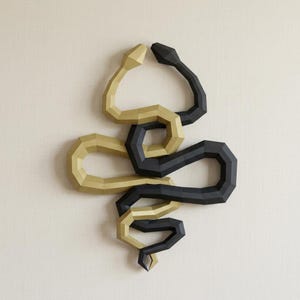 May include: A wall-mounted sculpture featuring two intertwined snakes. One snake is gold, the other is black. The snakes are stylized with geometric shapes and are positioned against a neutral background.