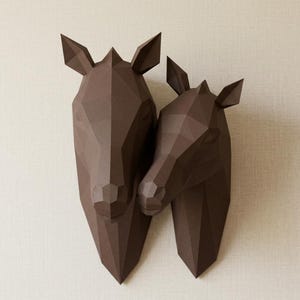 May include: Two brown geometric paper deer head wall decor. The larger head is positioned next to the smaller head, creating a loving embrace. The deer heads are made of folded paper and are mounted on a light beige wall.