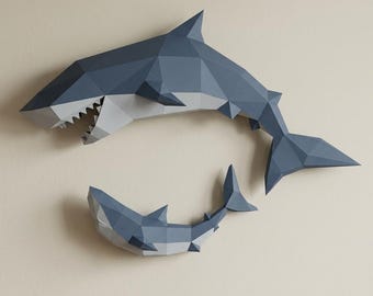 May include: Two geometric paper shark wall art pieces in shades of blue and gray. The larger shark is positioned above the smaller one, creating a dynamic composition. The sharks have sharp angles and detailed teeth.