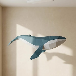 May include: A geometric paper whale sculpture in shades of blue, white, and grey, suspended from the ceiling. The whale's form is constructed from angular paper shapes, creating a modern, artistic aesthetic. The sculpture is a decorative item.