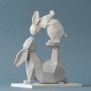 Op de afbeelding: Een geometrische papieren sculptuur van twee konijnen. Eén konijn zit op een voetstuk, de ander eronder, alsof ze elkaar aan het snuffelen zijn. De sculptuur is voornamelijk wit en grijs, tegen een blauwe achtergrond.