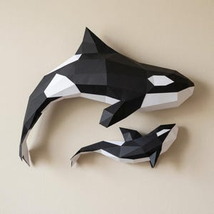 May include: Two papercraft orca whales, one large and one small, mounted on a beige wall. The whales are black and white, with geometric shapes creating their forms. The larger whale is positioned above the smaller one.