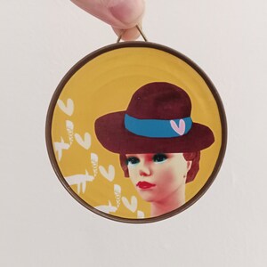 Puede incluir: Una lata redonda de color amarillo con un borde marrón, que presenta a una mujer con un sombrero marrón y una cinta azul. El sombrero tiene un corazón rosa en él. El fondo es amarillo con pájaros estilizados blancos.