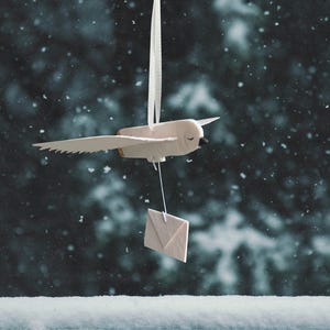 Könnte beinhalten: Ein weißer Vogel-Anhänger mit einem kleinen Umschlag, der daran hängt. Der Anhänger ist an einer weißen Schnur vor einem verschneiten Hintergrund befestigt.