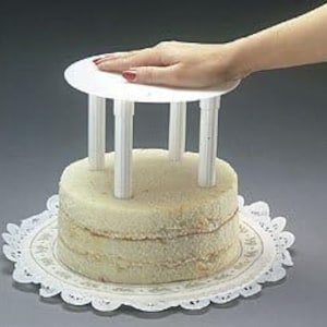 May include: White plastic cake stand with six support posts for stacking cakes. A hand is pressing down on the top of the stand.