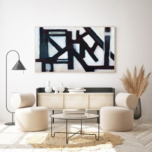 May include: Abstract black and white painting with geometric shapes on a white background, hanging above a modern living room with two beige armchairs, a coffee table, and a cabinet.