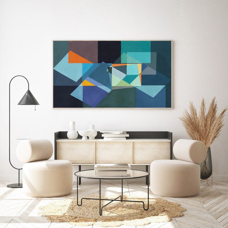 May include: Abstract geometric art print in a wooden frame. The artwork features overlapping shapes in shades of blue, teal, orange, and gray. The print hangs above a white cabinet with decorative items, two beige armchairs, and a round glass coffee table.
