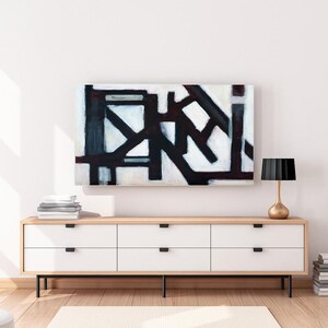 May include: A white dresser with six drawers and black handles sits on a wooden floor. A black and white abstract painting hangs above the dresser. A gold lamp with a black shade sits on the right side of the dresser.