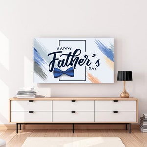 May include: A rectangular canvas print with the text "Happy Father's Day" and a blue bow tie illustration. The background has blue, grey, and orange brushstrokes. The print is displayed above a white cabinet with a wooden top and black legs.