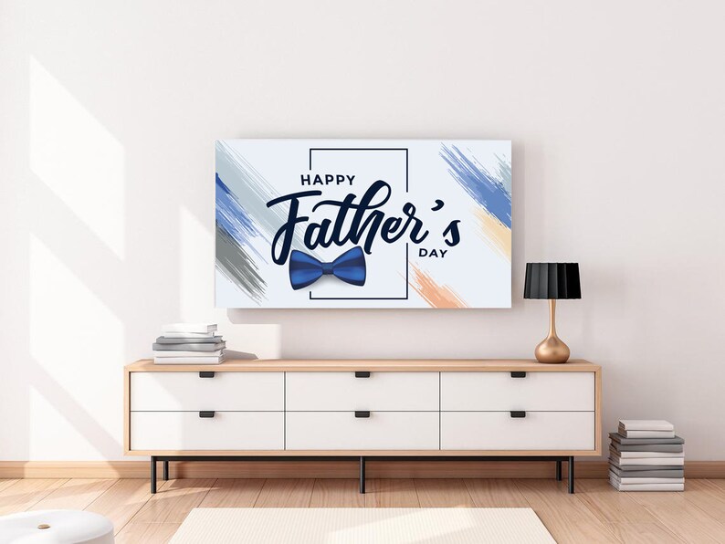 May include: A rectangular canvas print with the text "Happy Father's Day" and a blue bow tie illustration. The background has blue, grey, and orange brushstrokes. The print is displayed above a white cabinet with a wooden top and black legs.