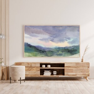 May include: Watercolor landscape art print featuring a serene mountain scene under a cloudy sky. The artwork showcases shades of blue, green, and purple, framed in a natural wood. The print is displayed above a wooden media console.