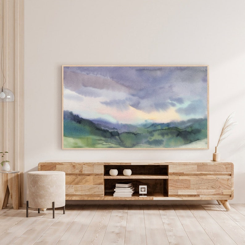 May include: Watercolor landscape art print featuring a serene mountain scene under a cloudy sky. The artwork showcases shades of blue, green, and purple, framed in a natural wood. The print is displayed above a wooden media console.