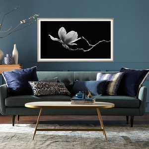 May include: A teal blue sofa with navy blue pillows and a white and black framed print of a magnolia flower on a black background. The sofa is in front of a teal blue wall and a coffee table with a marble top and gold legs.