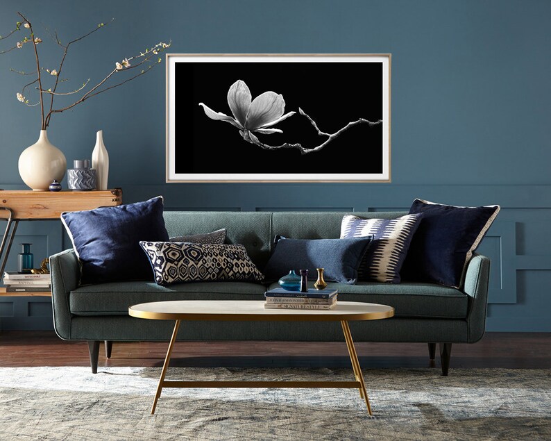 May include: A teal blue sofa with navy blue pillows and a white and black framed print of a magnolia flower on a black background. The sofa is in front of a teal blue wall and a coffee table with a marble top and gold legs.