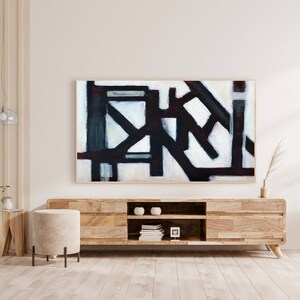 May include: Abstract painting with black, white, and gray geometric shapes on a white background. The painting is framed in a natural wood frame and is hanging on a white wall above a wooden media console.