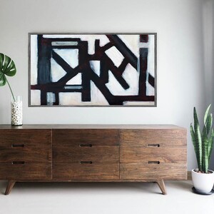 May include: A dark wood dresser with six drawers and a modern design. The dresser is in front of a white wall with a black and white abstract painting hanging above it. A green plant is on the right side of the dresser.