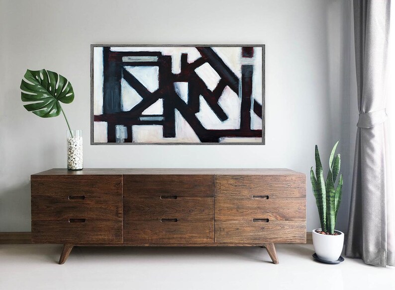 May include: A dark wood dresser with six drawers and a modern design. The dresser is in front of a white wall with a black and white abstract painting hanging above it. A green plant is on the right side of the dresser.