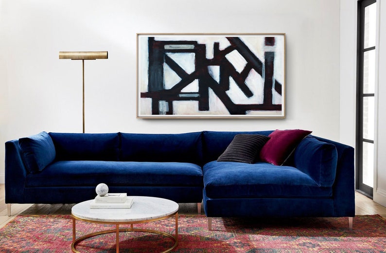 May include: A blue velvet sectional sofa with a black and white abstract painting above it. The sofa has two throw pillows, one black and one burgundy. A gold floor lamp is on the left side of the sofa. A round marble coffee table sits in front of the sofa.