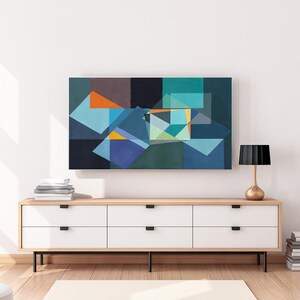 May include: An abstract geometric painting in shades of blue, orange, and green hangs above a white and wood-tone cabinet. A gold lamp with a black shade sits on the cabinet, next to a stack of books. The room has a modern, minimalist aesthetic.