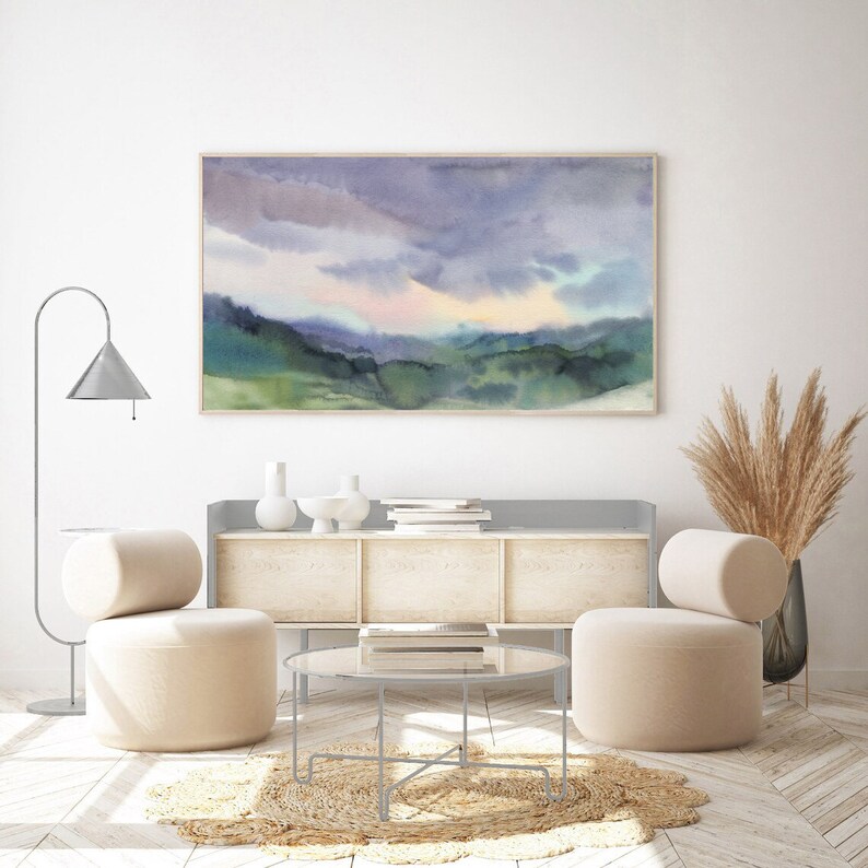 May include: A framed watercolor landscape painting with a serene sky and rolling hills in shades of blue, green, and purple. The artwork hangs above a modern living room setup with neutral-toned furniture and decor.