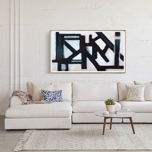 May include: Abstract art print in a white frame with black and white geometric shapes and a red accent. The print is hanging on a white brick wall above a white sectional sofa with pillows.