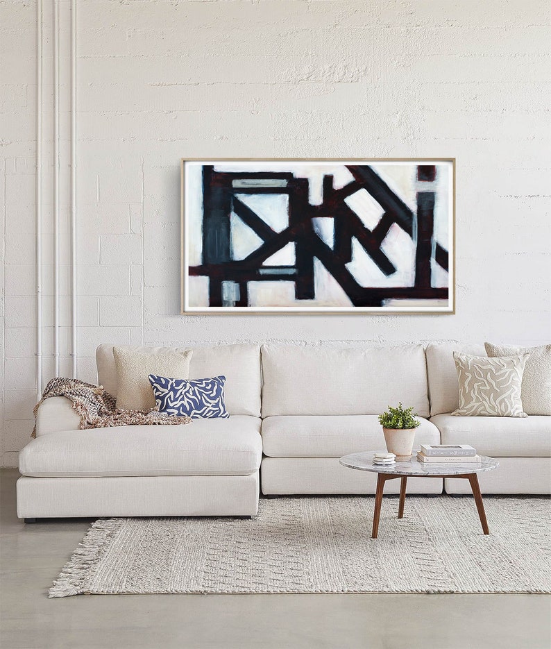 May include: Abstract art print in a white frame with black and white geometric shapes and a red accent. The print is hanging on a white brick wall above a white sectional sofa with pillows.