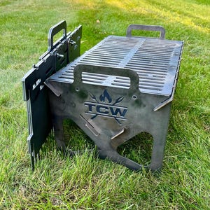 May include: A black metal portable grill with a rectangular cooking surface and side panels. The grill has handles and the text "TCW" with a flame and spear design. It is set on a grassy surface.