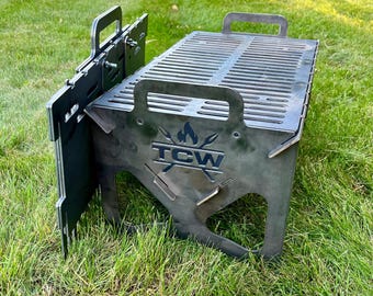 Pode incluir: Uma grelha portátil de metal preto com uma superfície de cozedura retangular e painéis laterais. A grelha tem pegas e o texto "TCW" com um design de chama e lança. Está colocada sobre uma superfície relvada.