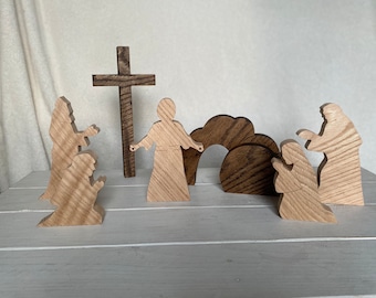 May include: Wooden figurines depicting the Easter story. The figures include Jesus, two women, and a tomb. The figures are made of light brown wood and are standing on a white surface.