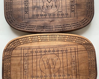 May include: Two wooden cribbage boards with football field designs. One board features the University of Michigan logo and the other features the Washington Huskies logo. Both boards have a dark brown finish and are oval-shaped with drilled holes.