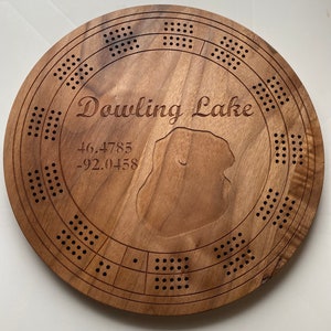 May include: A wooden cribbage board with a circular shape and a cutout of a lake. The board is engraved with the text "Dowling Lake" and the coordinates "46.4783 -92.0458".