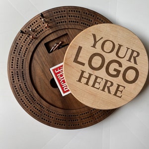May include: A wooden cribbage board with a walnut finish and a removable lid. The lid is made of maple wood and has the text "YOUR LOGO HERE" engraved on it.