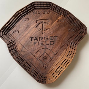 May include: A wooden cribbage board shaped like a baseball field with the words "TARGET FIELD" and the Minnesota Twins logo. The board has a dark brown stain and is made of walnut wood.