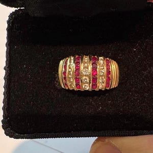 May include: A gold ring featuring alternating vertical channels of round red rubies and clear white diamonds. The ring has a wide, domed band design with ribbed gold detailing on the outer edges, presented inside a black velvet jewelry box.