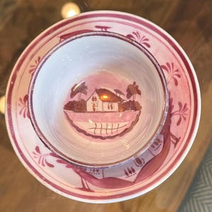 May include: A ceramic teacup and saucer set featuring a white base with pink hand-painted details. The interior of the cup and the saucer display a landscape illustration of a house with trees and a fence, rendered in shades of pink and brown.