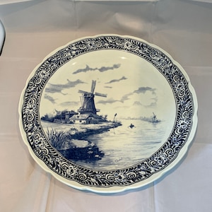 May include: A round ceramic plate with a scalloped edge featuring a blue and white landscape illustration of a windmill by a river. The border is decorated with a repeating floral and scroll pattern. The artist signature is visible near the water.