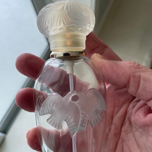 May include: Clear glass perfume bottle with a frosted, decorative dove design. The bottle has a frosted glass stopper with a floral pattern and a gold-colored metal ring around the neck.