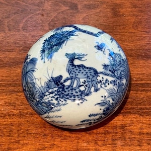 May include: A round ceramic lidded box featuring a blue and white hand-painted design. The lid displays a detailed scene of two spotted deer standing and resting among stylized trees, foliage, and small insects on a pale off-white background.