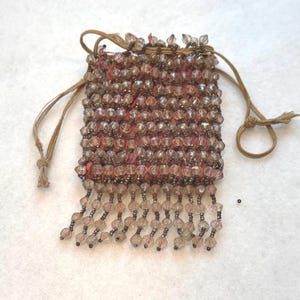 May include: A small rectangular drawstring pouch crafted from a pink fabric base, entirely covered in rows of faceted, translucent, light-brown beads. The bottom edge features a fringe of dangling beaded strands, and two thin tan cords serve as closures.