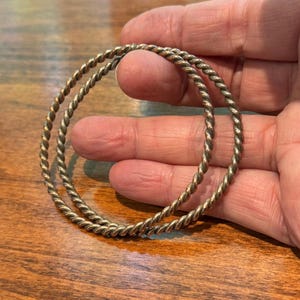 May include: Two circular bangles made of a metallic material with a twisted, rope-like texture. The bangles have a silver-toned finish with subtle darkened areas within the grooves of the twisted design, creating a consistent, uniform appearance.